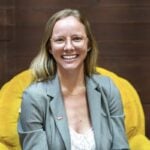Kristin Sullivan, blonde with glasses, gray blazer, and necklace, smiles on a yellow chair before a wooden wall at Loqal Marketing.