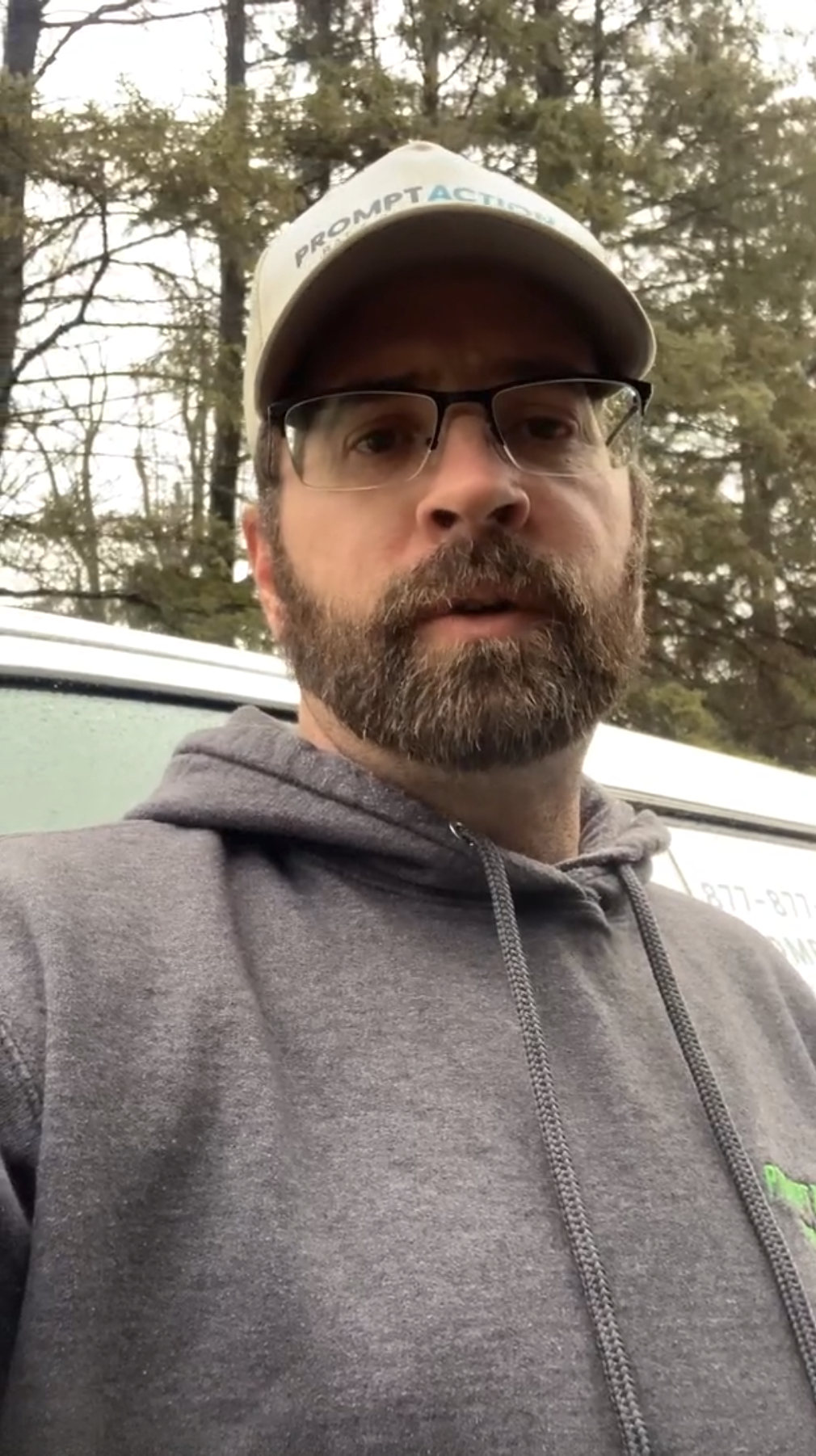 A bearded man in glasses, a cap, and a gray hoodie stands outside in front of a white vehicle and trees, representing Loqal Marketing’s approachable team.