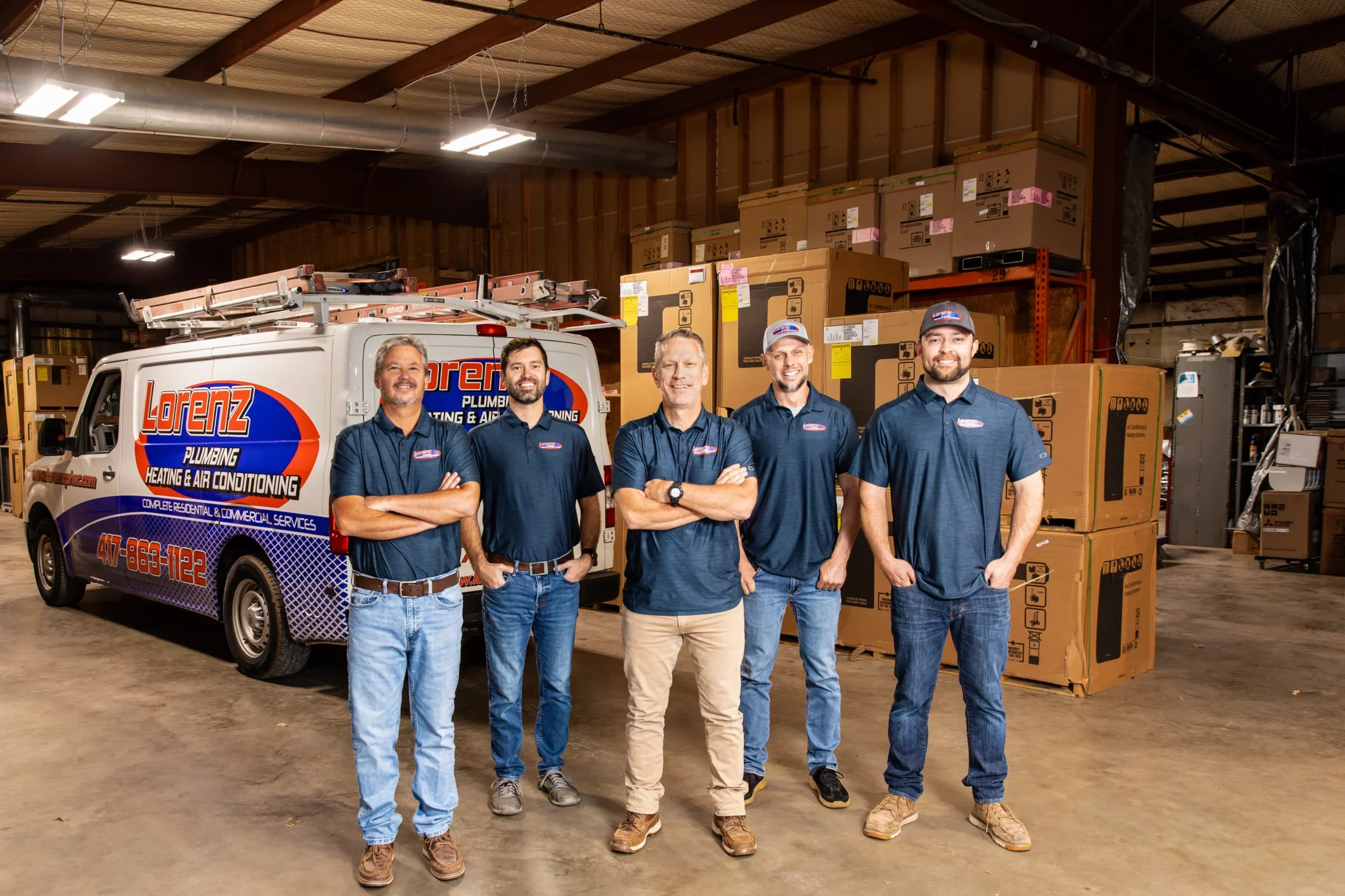 Five uniformed team members pose in a warehouse among HVAC boxes, standing in front of a branded service van for a plumbing and heating company specializing in air conditioning solutions—a professional crew ready to meet your service needs.