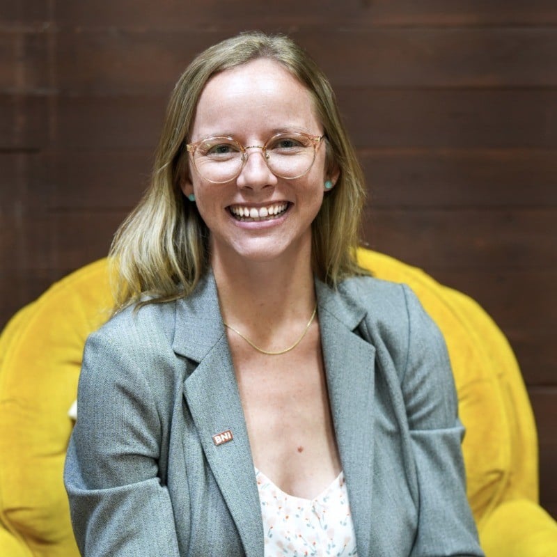 Kristin Sullivan, blonde with glasses, gray blazer, and necklace, smiles on a yellow chair before a wooden wall at Loqal Marketing.