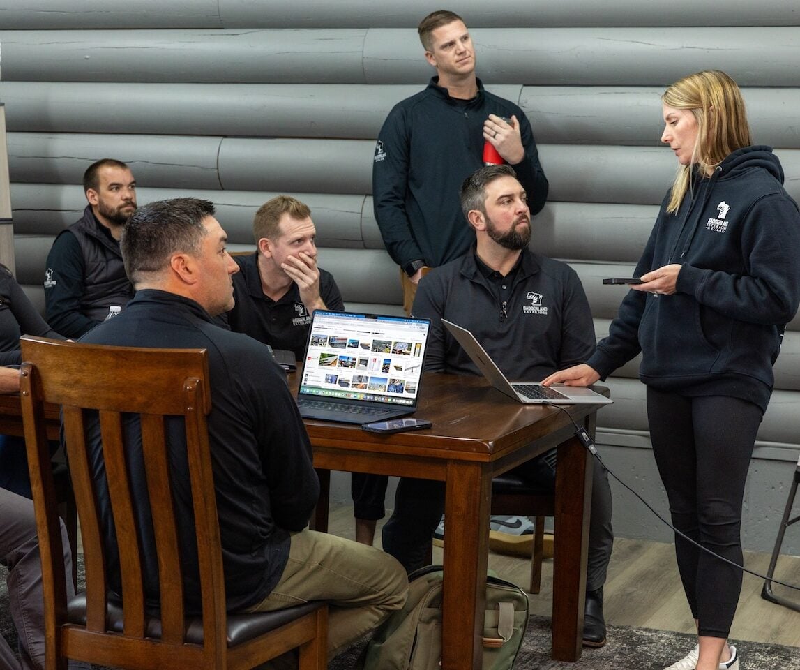A team of seven in matching black sweatshirts collaborates around a wooden table with open laptops, actively discussing roofing and siding strategies in a modern, gray log-walled room. A woman stands at the front, presenting with a tablet to the group at Badgerland Exteriors.