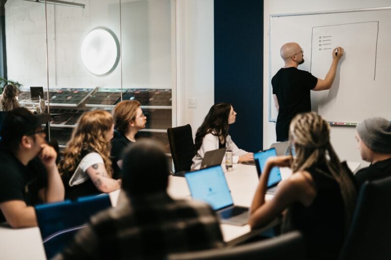 A team in a modern office focuses as one member writes key points on a whiteboard; others, equipped with laptops, engage attentively at the conference table.