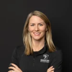 Steph Laurer of Baggerland Exteriors stands confidently with arms crossed, smiling, in branded attire against a black background.